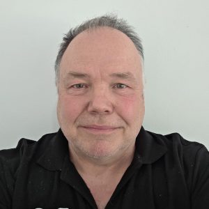 Selfie of a smiling middle-aged man with short gray hair, wearing a black polo with an orange logo, against a pale wall.