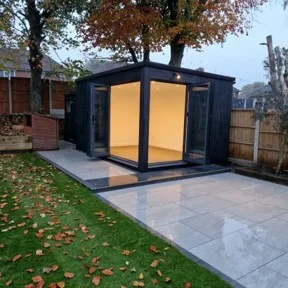 Modern glass garden room with a black metal frame, illuminated interior, set on a tiled patio of a backyard with fall leaves nearby.