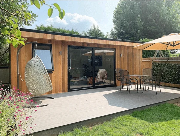 Wooden cabin with sliding glass doors opening to a wooden deck, outdoor dining set with a beige umbrella; a hanging chair on the left and a green lawn beyond.